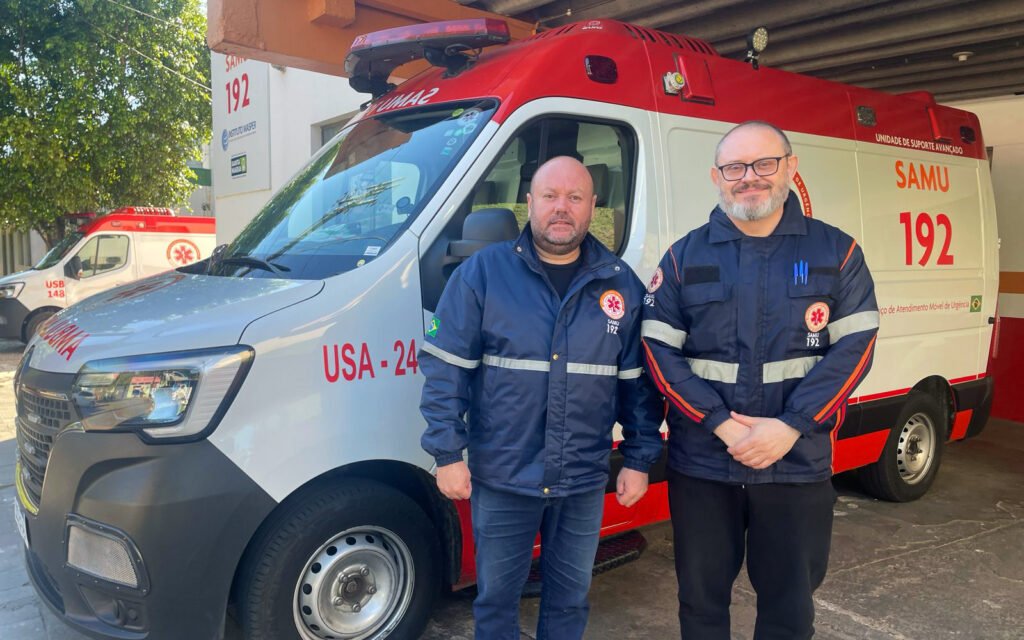 Dois profissionais do SAMU (Serviço de Atendimento Móvel de Urgência) posam em frente a uma ambulância de suporte avançado na base do SAMU em Santa Maria (RS). Ambos usam uniformes do serviço, e ao fundo há outra ambulância estacionada. A imagem destaca a estrutura e a equipe dedicada ao atendimento pré-hospitalar emergencial.